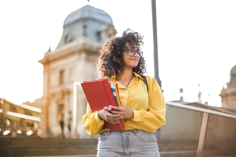 Student in yellow shirt holding folders outside university building at golden hour