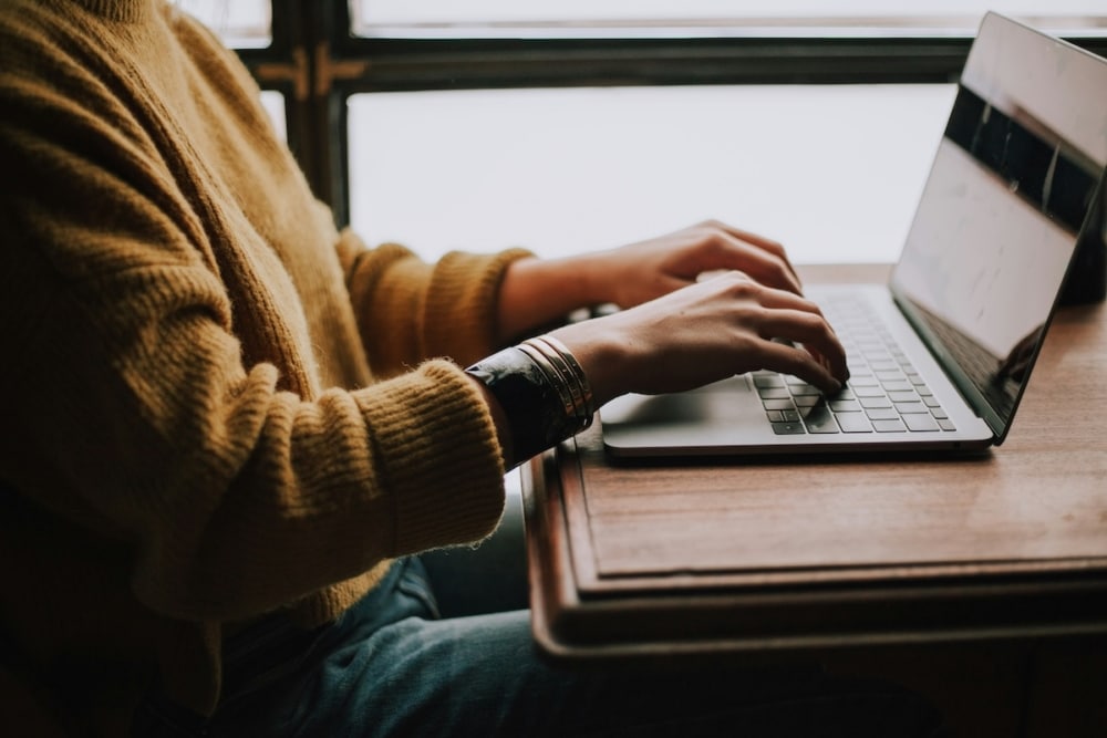 A person typing on a laptop at a wooden desk near a window.