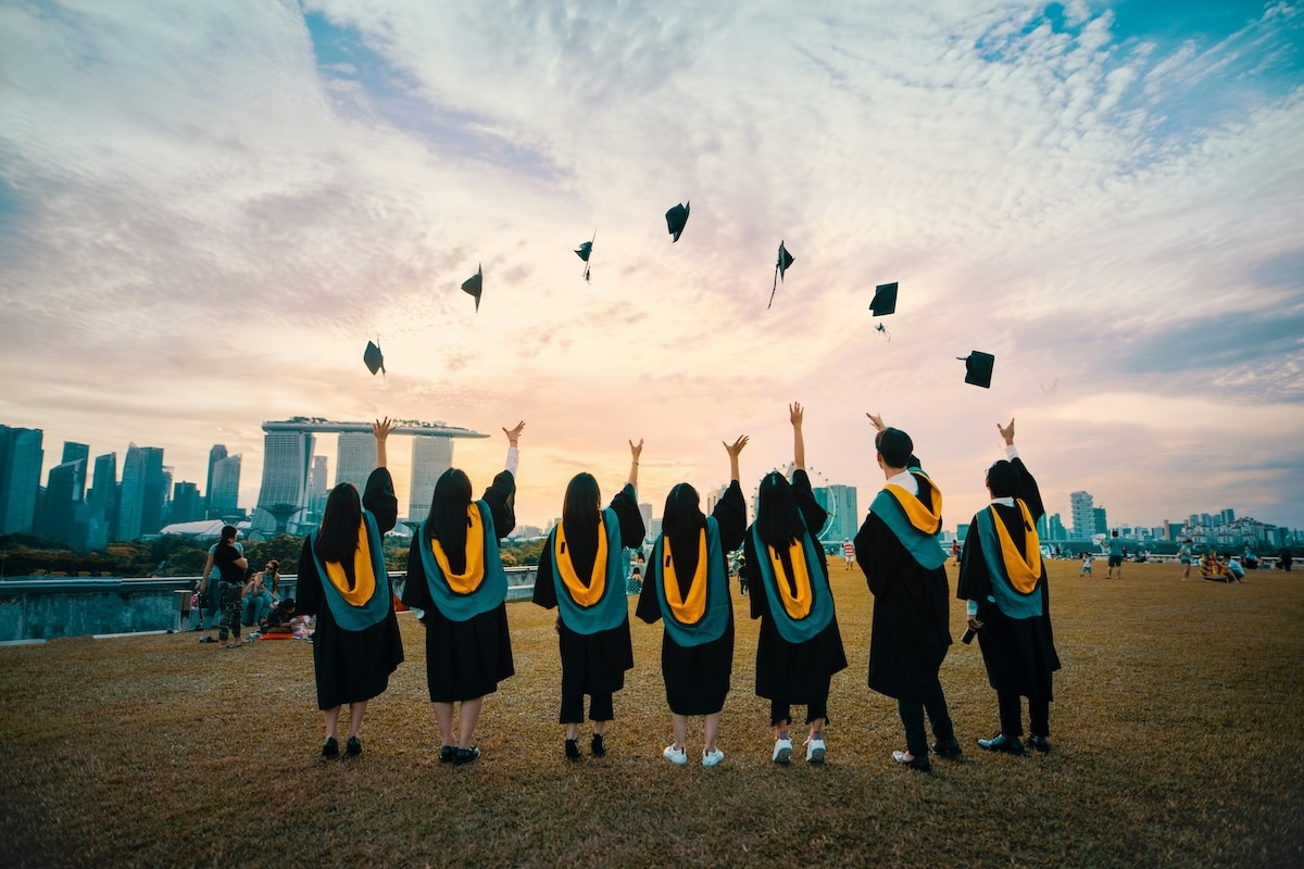 Graduates in caps and gowns celebrating by throwing their hats into the air at sunset.