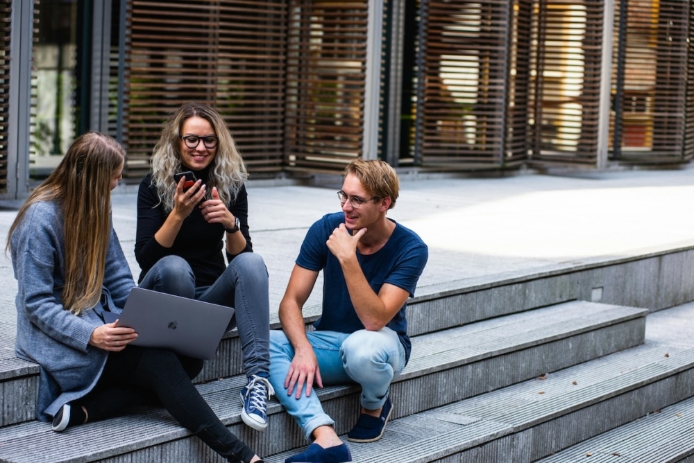 Three students sitting on outdoor steps, smiling and using a phone and laptop together.