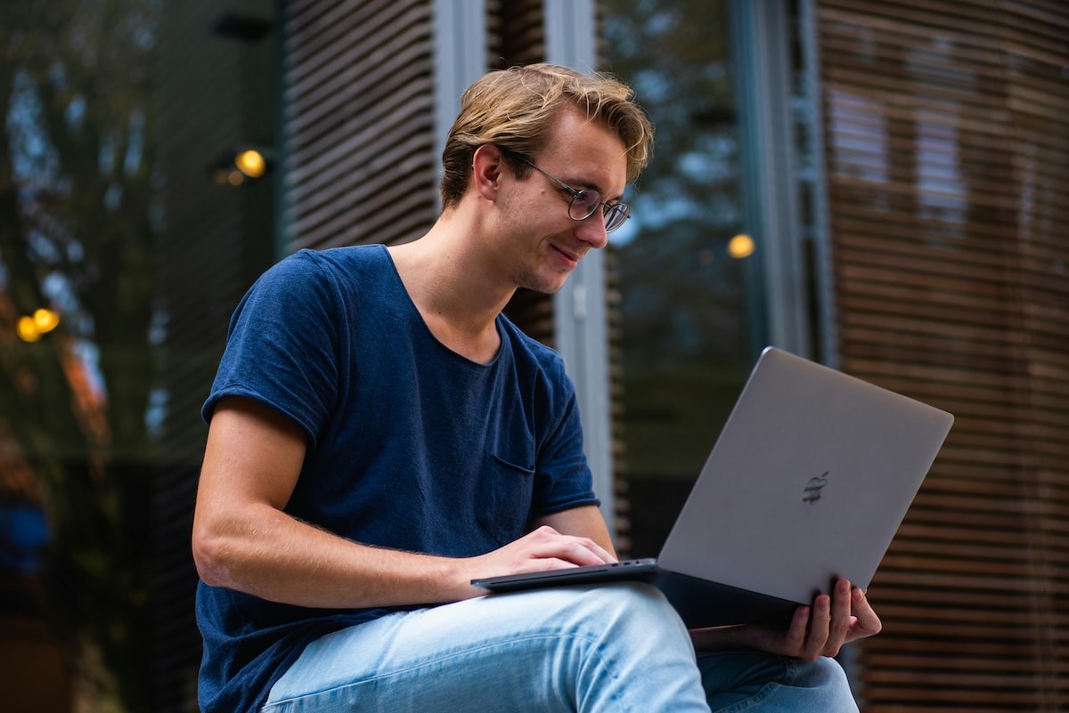 A young man sitting outdoors working on a laptop.