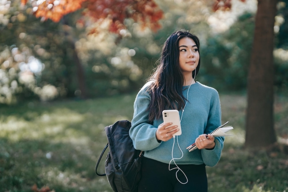 Student standing outdoors holding phone, notebook, and pen with backpack on shoulder
