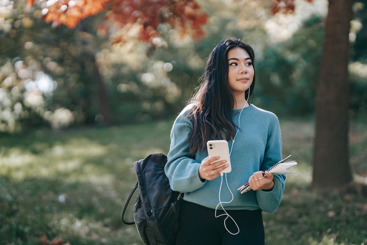Student standing outdoors holding phone, notebook, and pen with backpack on shoulder