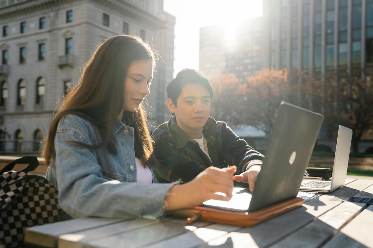 Two college students studying outdoors at a wooden table with laptops open in front of them.