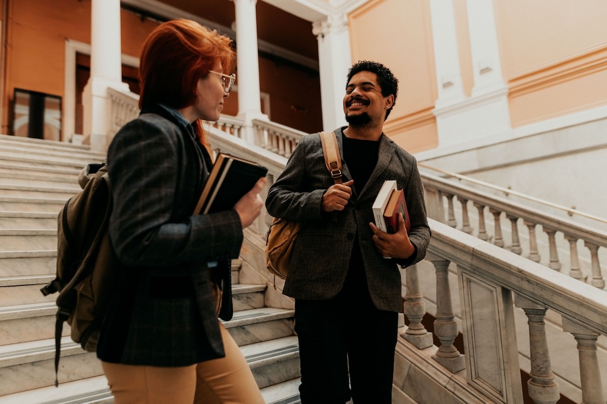 Two college students with backpacks and books having a conversation on a marble campus staircase.