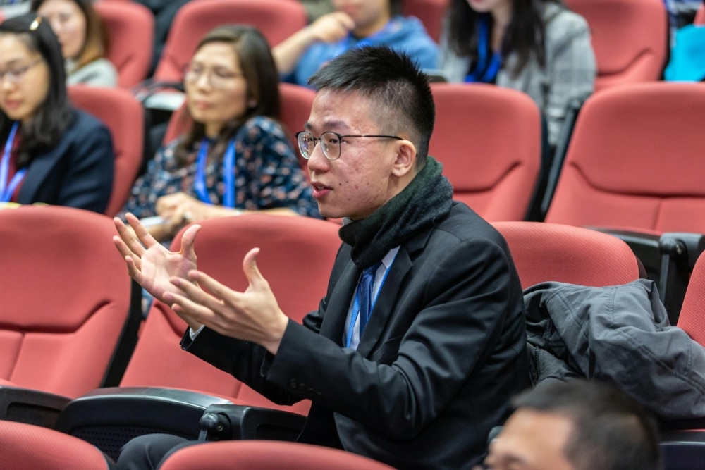 A man wearing a black jacket sits in a lecture hall.