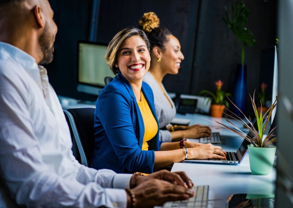 Positive Workplace Collaboration in Tech Environment Smiling businesswoman in blue blazer working at a computer with colleagues.