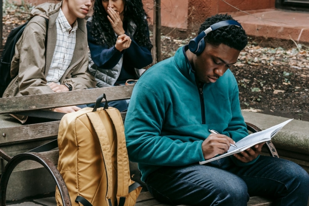 Student sitting on a park bench writing in a notebook while wearing headphones
