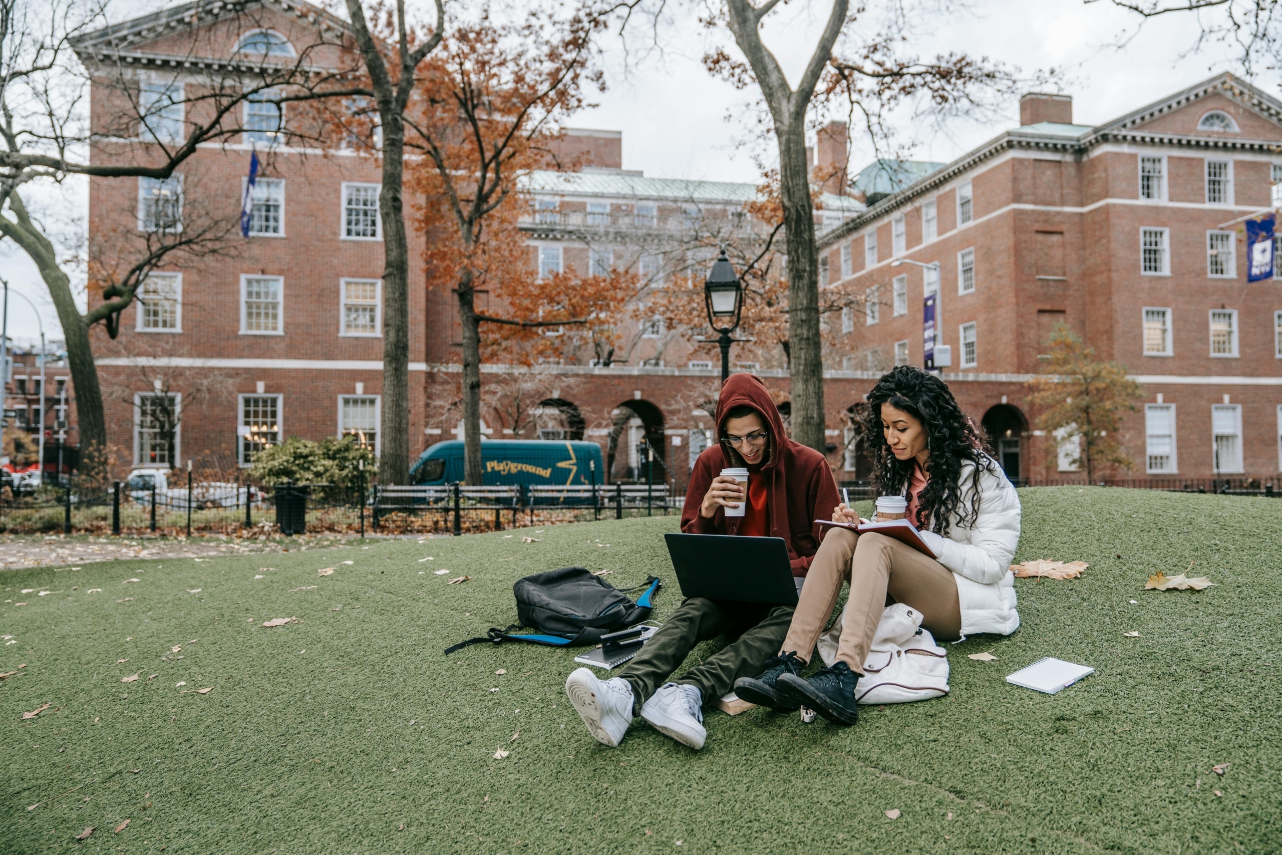 Two college students sitting on campus lawn using a laptop together in front of red brick university buildings