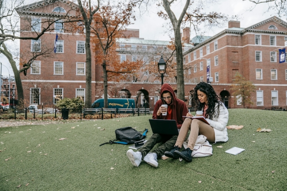 Two students studying outdoors on campus with a laptop and notebooks in autumn.