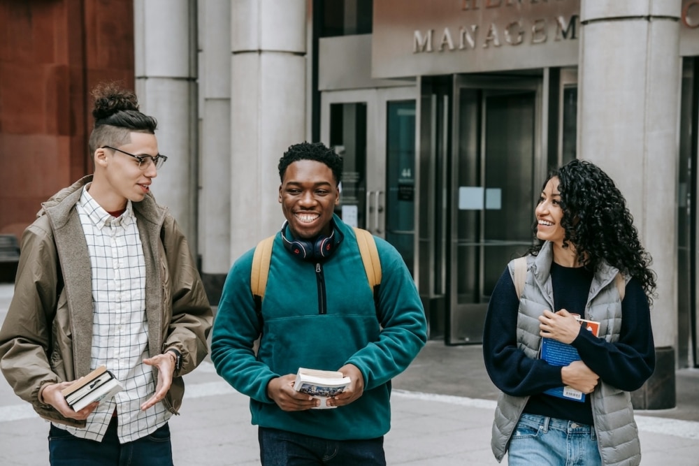 Three college students walking together outside a university building, smiling and carrying books.