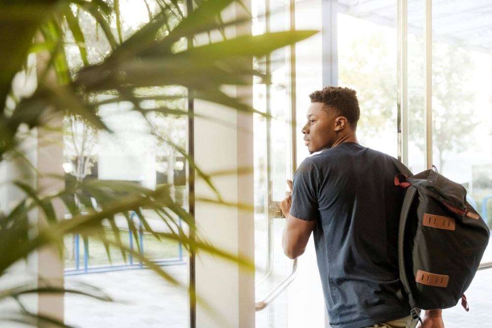 black young man with a backpack on his right shoulder is walking through the glass door