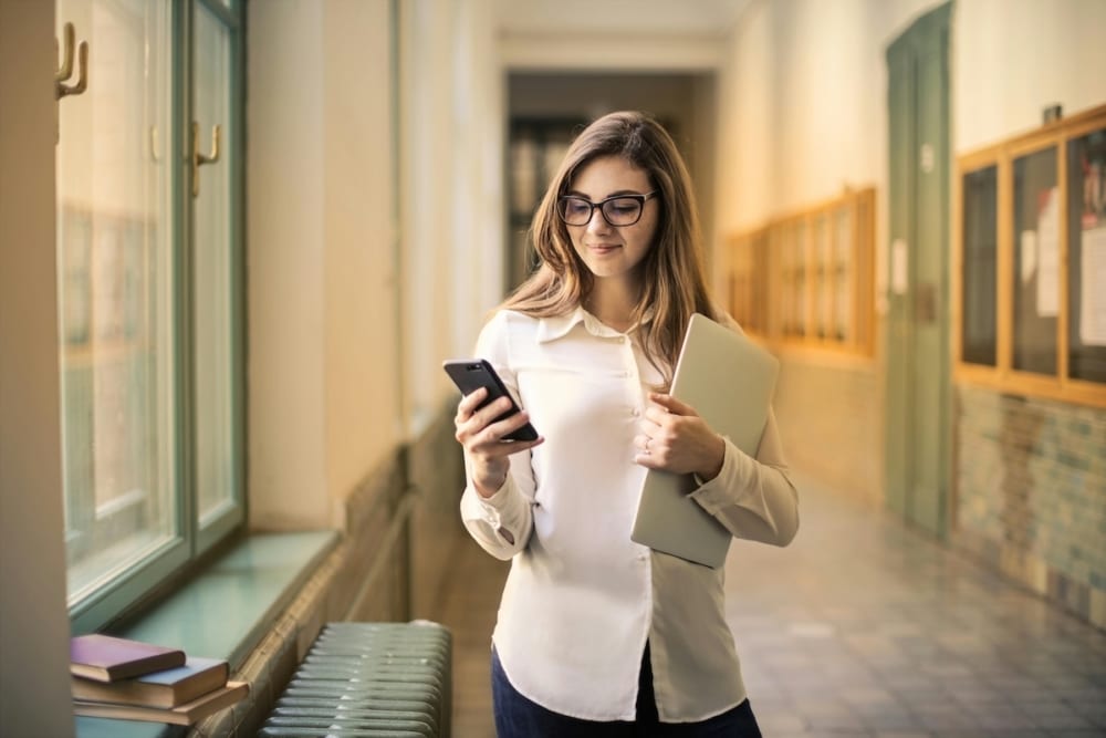 A woman in a hallway holding a laptop and checking her smartphone.