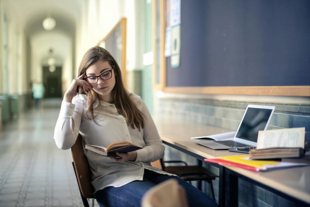 A young woman is sitting in at the workplace equipped in a hallway and reading a book