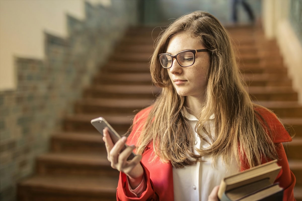 Student Checking Phone Between Classes Young woman holding books and using her smartphone while standing near stairs inside a school building.