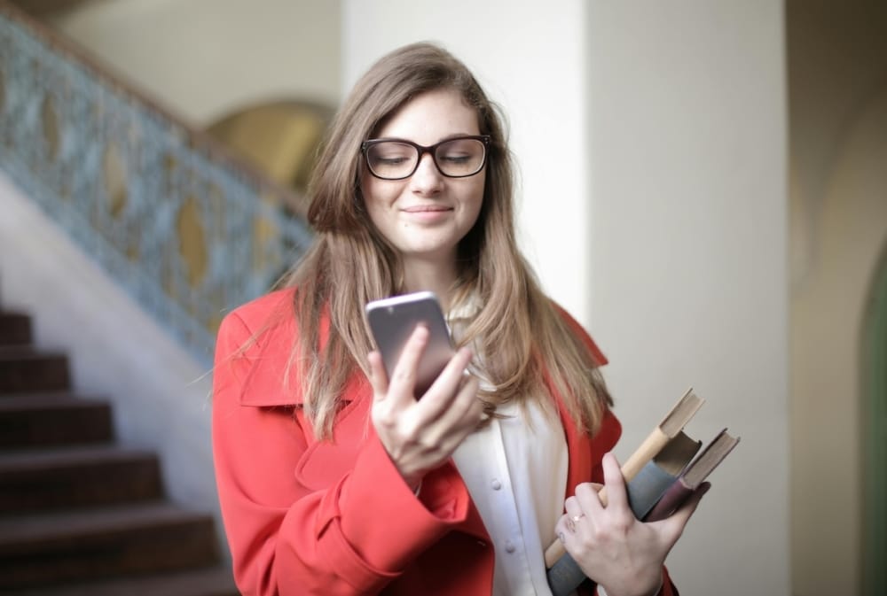 A young woman in a red coat holding books and checking her smartphone.