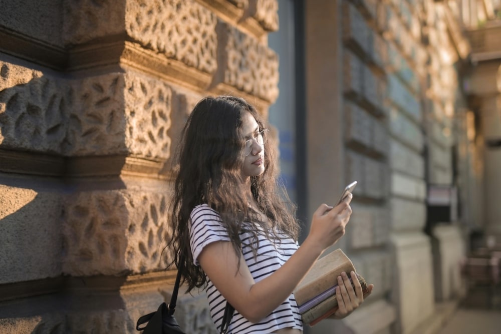 Young woman with long hair holding books and checking her phone outdoors