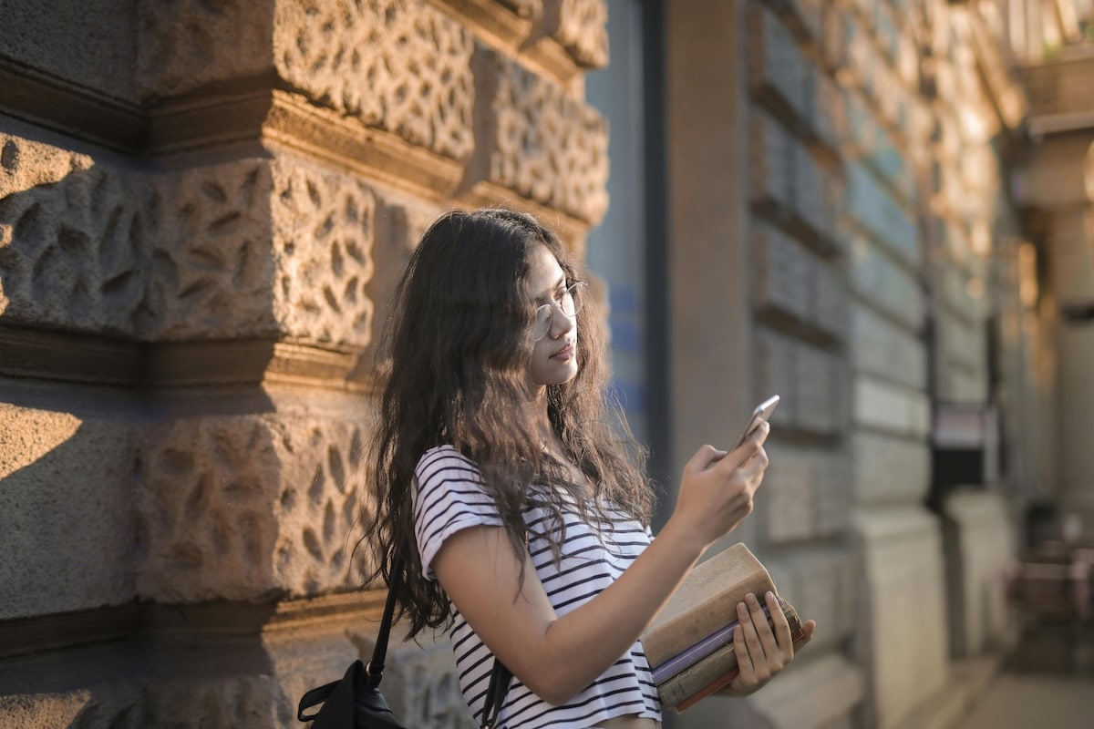 Student Using Phone Outside a Historic Building Young woman with long hair holding books and checking her phone outdoors
