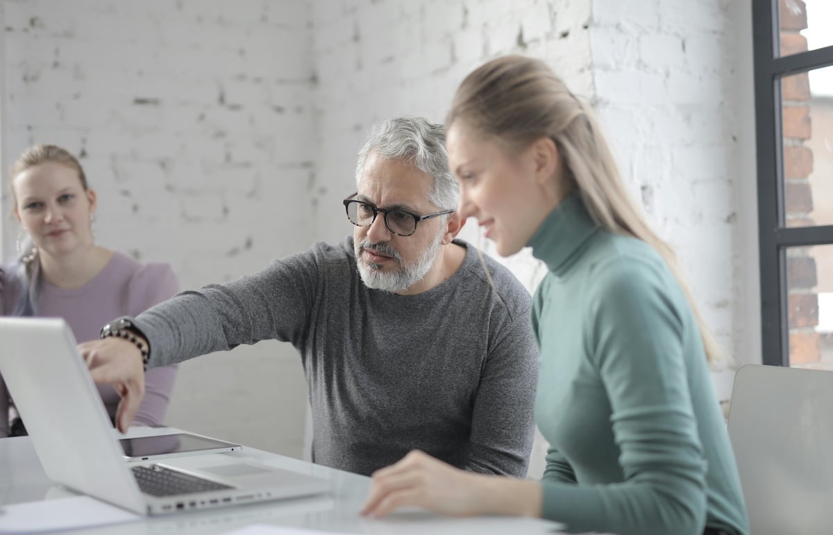 Older male mentor assisting a younger female colleague with work on a laptop in a bright office