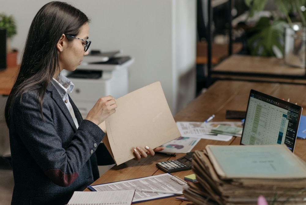 Admissions counselor reviewing student files at her desk, with a stack of folders and a data dashboard visible on her laptop screen.