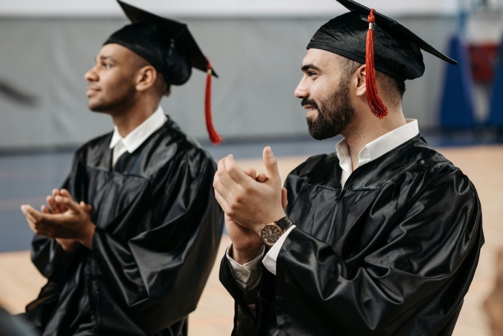 Two male graduates in black caps and gowns clapping during a commencement ceremony