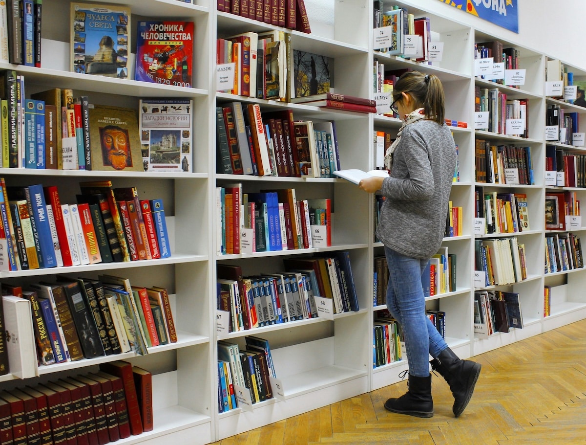 A woman reading a book in a library surrounded by shelves filled with books.