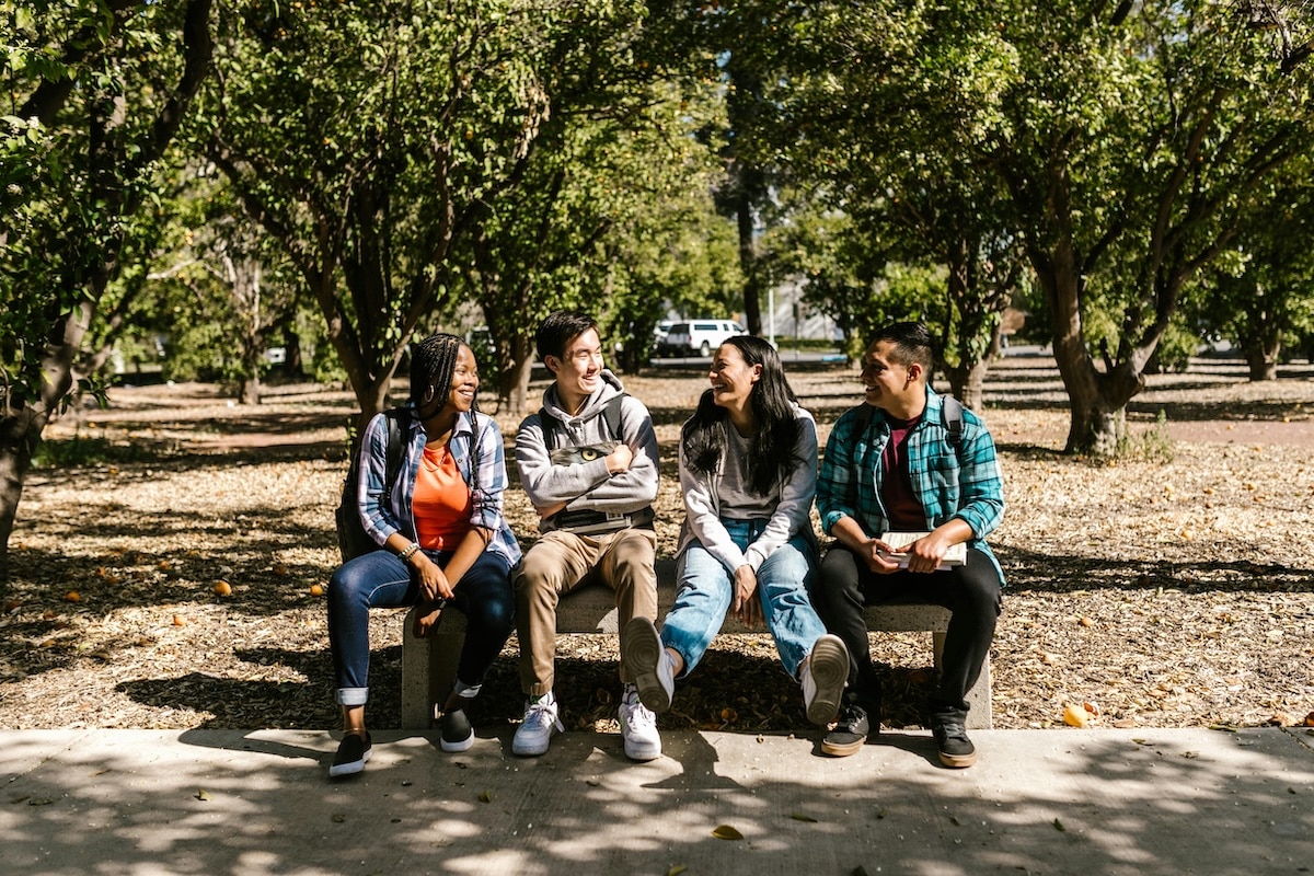 Four diverse college students sitting on a bench outdoors, chatting and laughing under the trees