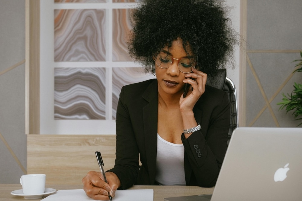 Professional woman in glasses on phone call while writing notes at her desk