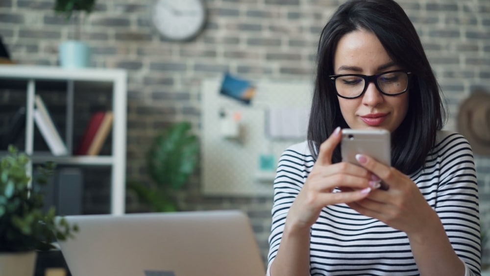 Woman with glasses checking her phone at a desk with a laptop open in front of her