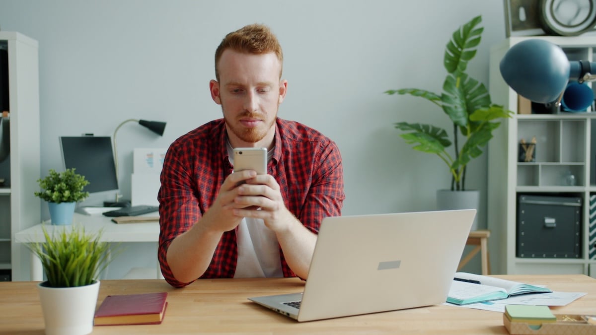 Man checking his phone at a desk with a laptop open in front of him