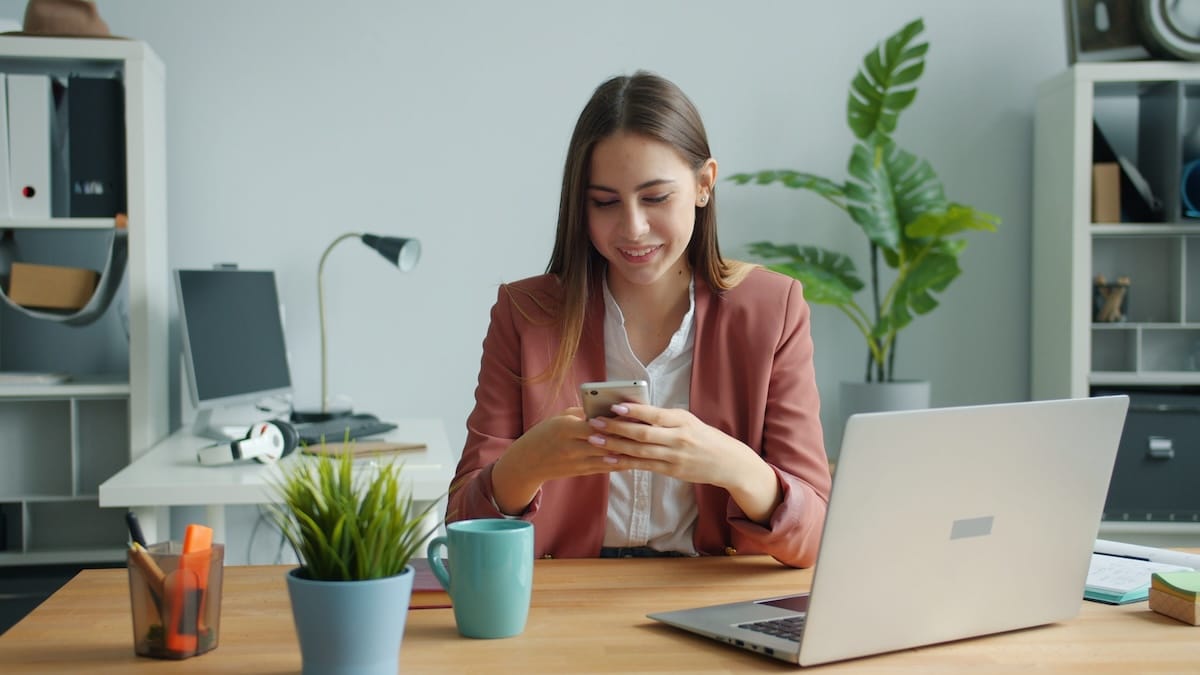 Smiling higher ed staff member texting on her phone at her desk, with a laptop, notebook, and plants nearby