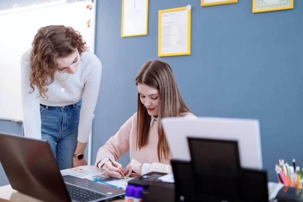 Two young women collaborating in a classroom, one seated and writing while the other leans over to assist