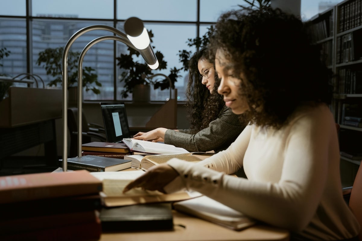 Two women reading and highlighting books under a library desk lamp