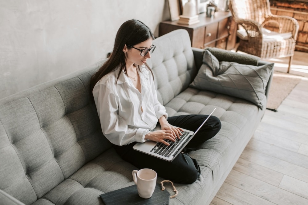 Woman working on laptop while sitting cross-legged on a grey sofa