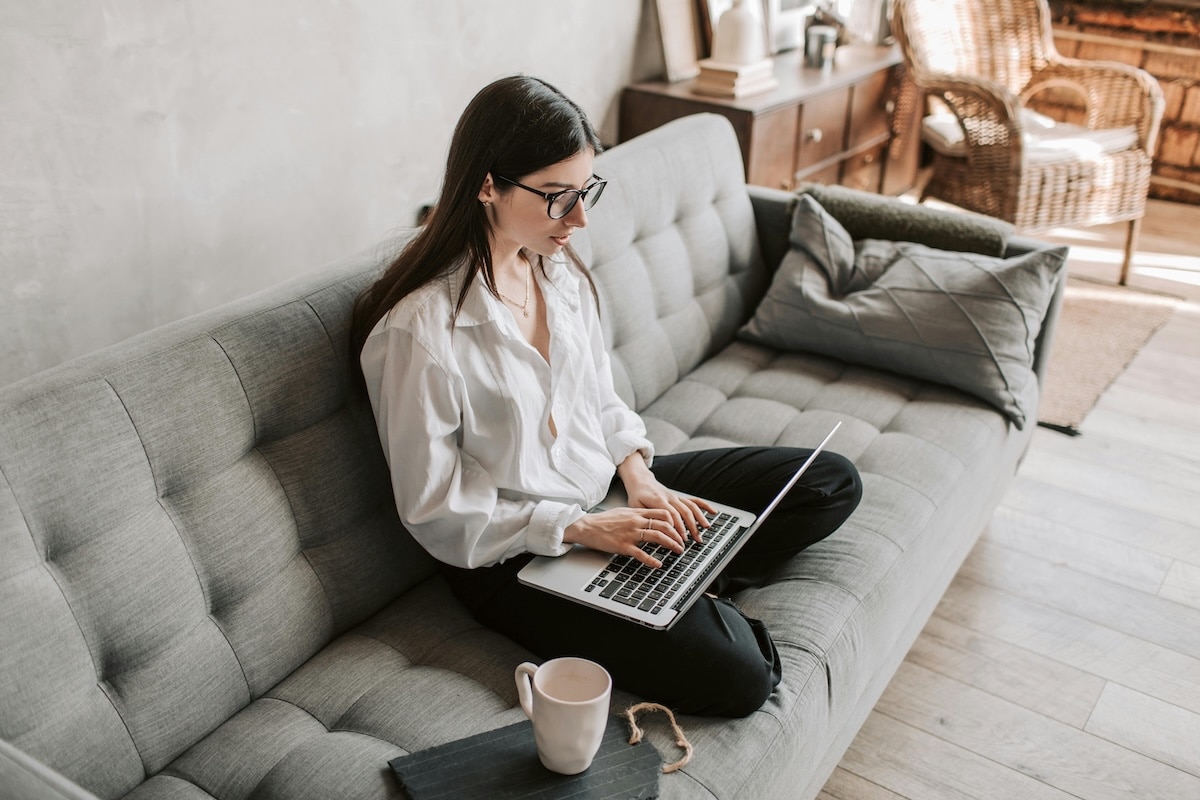 Woman working on laptop while sitting cross-legged on a grey sofa