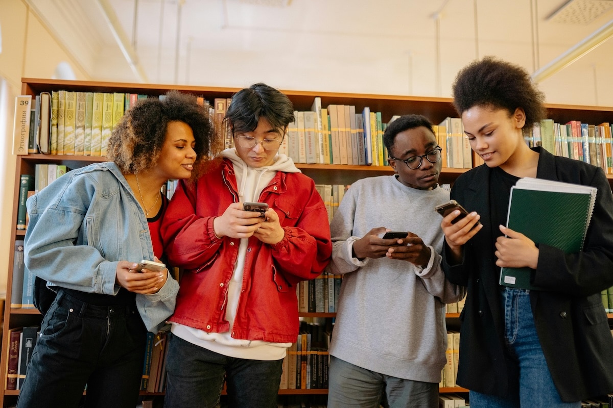 Group of diverse college students standing in front of library bookshelves, looking at their smartphones together