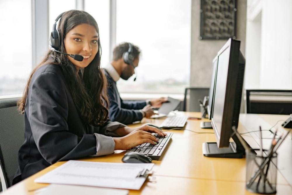 Woman with headset smiling while working at a desktop computer.