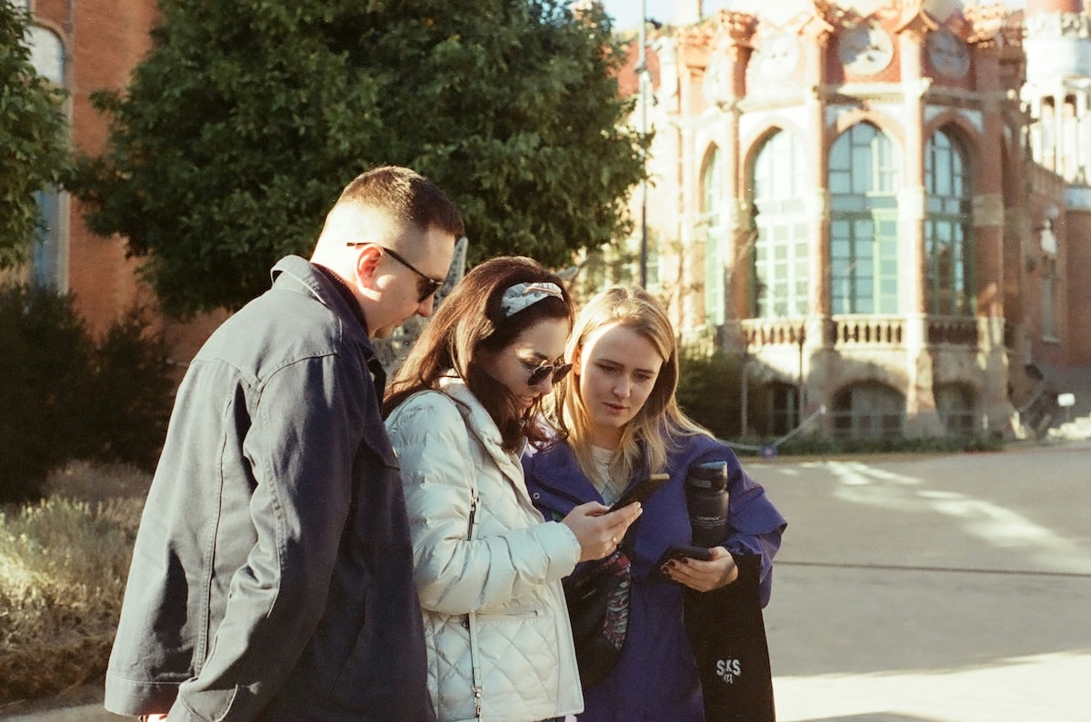 Three adults standing outdoors in front of a historic brick building, looking at a phone screen together.