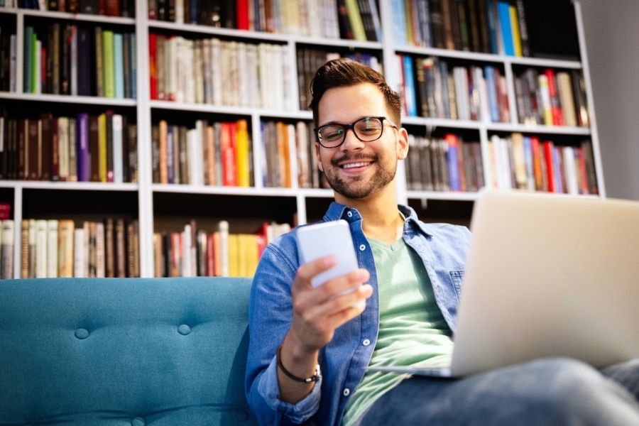 Smiling man sitting on a sofa in front of bookshelves, using a smartphone and laptop.