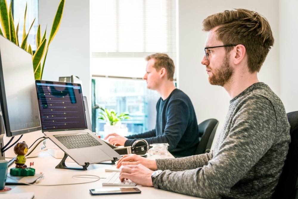 Two focused male programmers working at modern office desks with multiple monitors and laptops