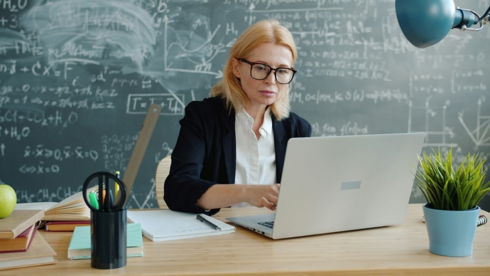 Female educator working on a laptop in front of a chalkboard filled with mathematical equations