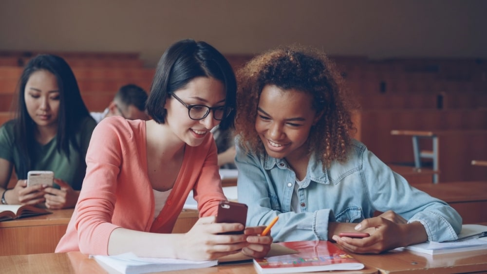 Two students smiling while looking at a smartphone together in a classroom